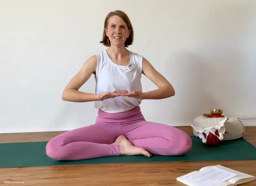 Eine Frau in pinken Yogaleggings sitzt auf einer Yogamatte und erklärt etwas.