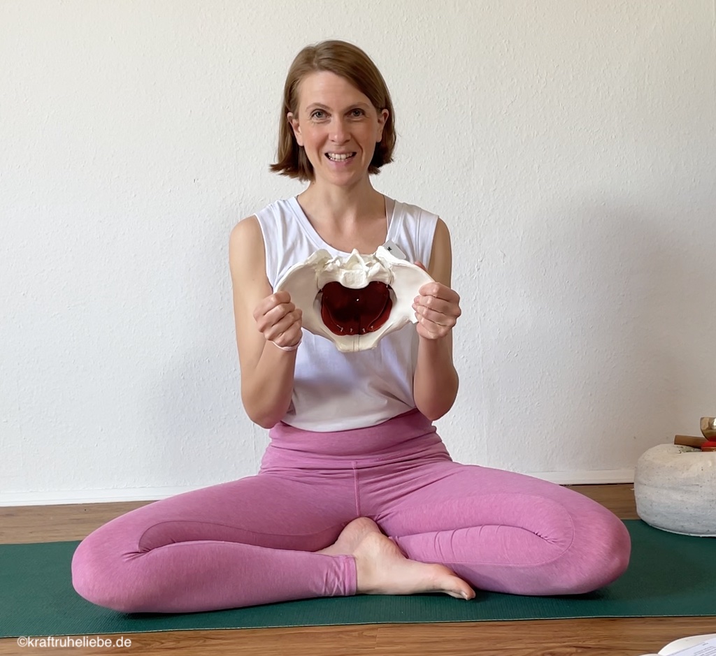 Eine Yogalehrerin sitzt auf einer Yogamatte und hält ein Modell eines weiblichen Beckens in den Händen
