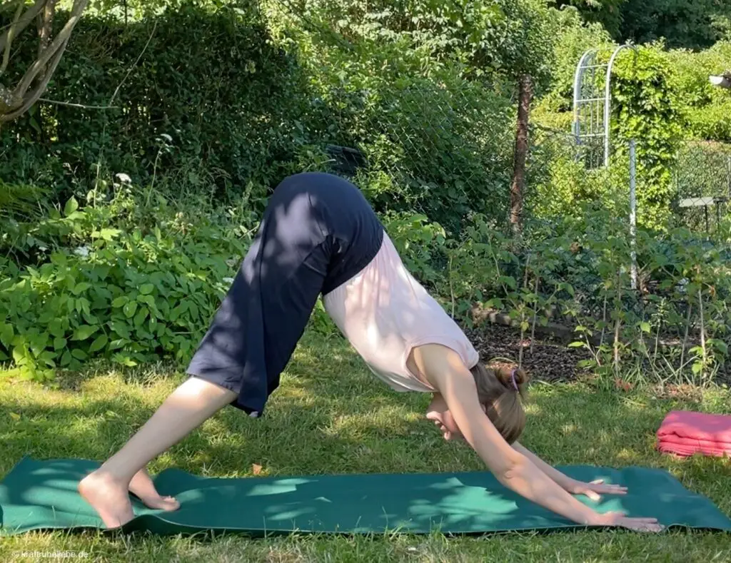 Eine Frau in Yogakleidung übt die Hundestellung auf einer grünen Yogamatte in einem Garten.