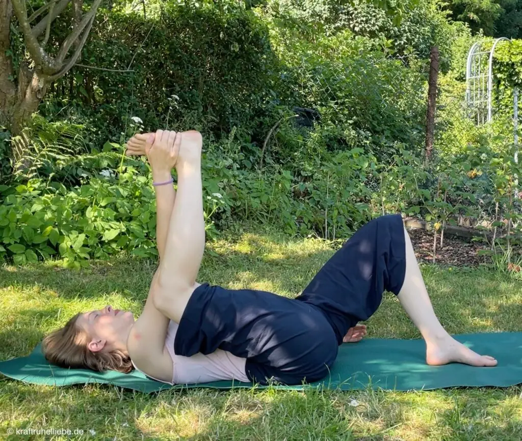 Eine Frau übt halbbeinig Ananda Balasana auf einer Yogamatte im Garten