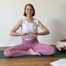 Eine Frau in pinken Yogaleggings sitzt auf einer Yogamatte und erklärt etwas.