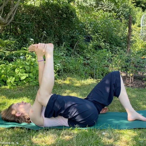 Eine Frau übt halbbeinig Ananda Balasana auf einer Yogamatte im Garten